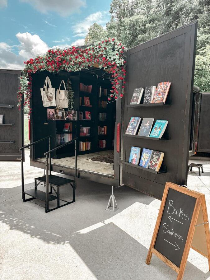She's Behind Nashville’s First Romance-Only Bookstore - 3 A black mobile bookshop in Nashville with an open door displays shelves of books, tote bags, and flowers. An easel sign reads "Books > Sadness" with an arrow pointing toward the inviting local shop.