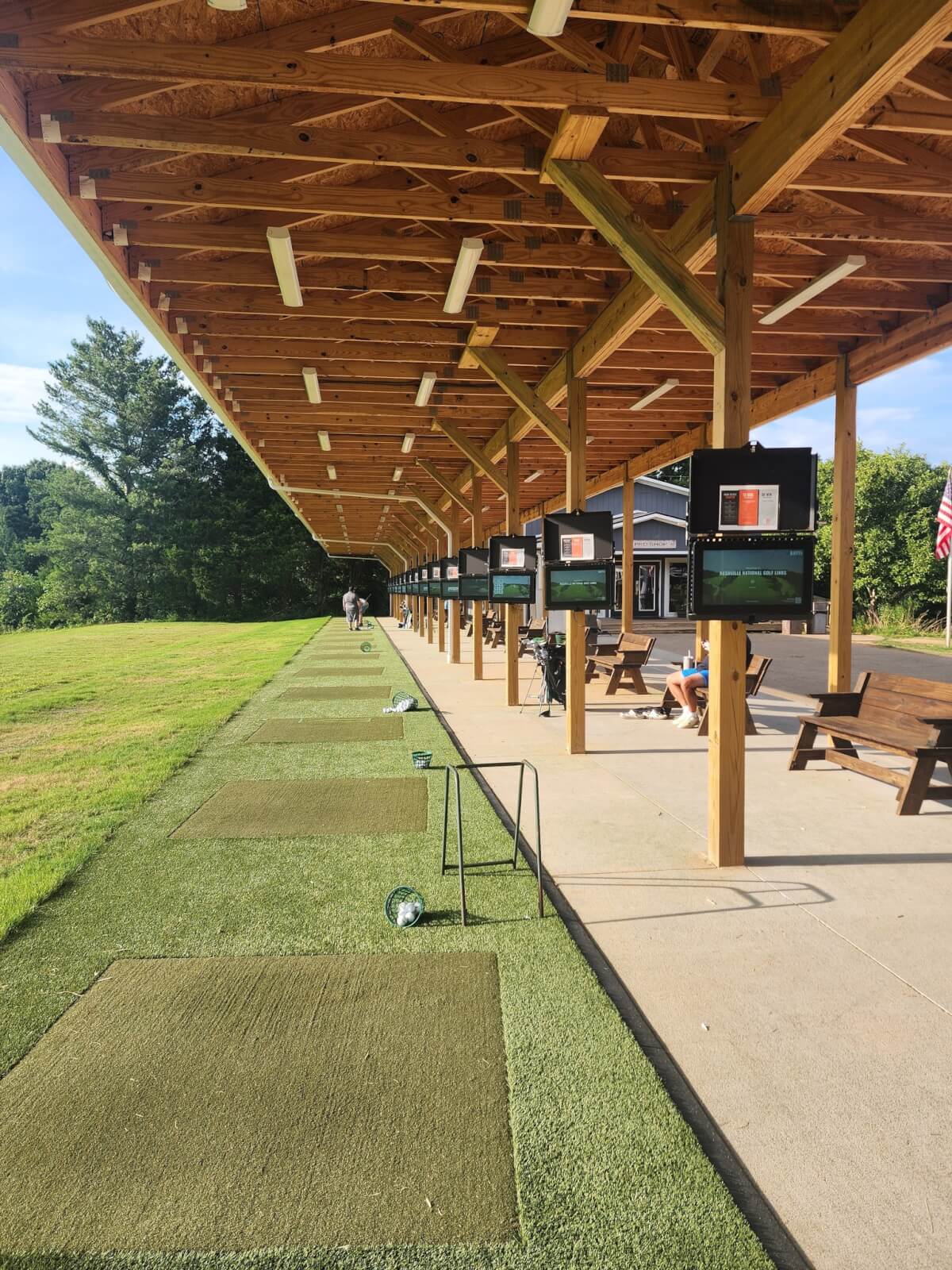 Covered golf driving range with artificial turf mats, overhead monitors, and people practicing their golf swings on a sunny day. Trees and a building are visible in the background, offering a relaxed vibe found at top Nashville golf courses.