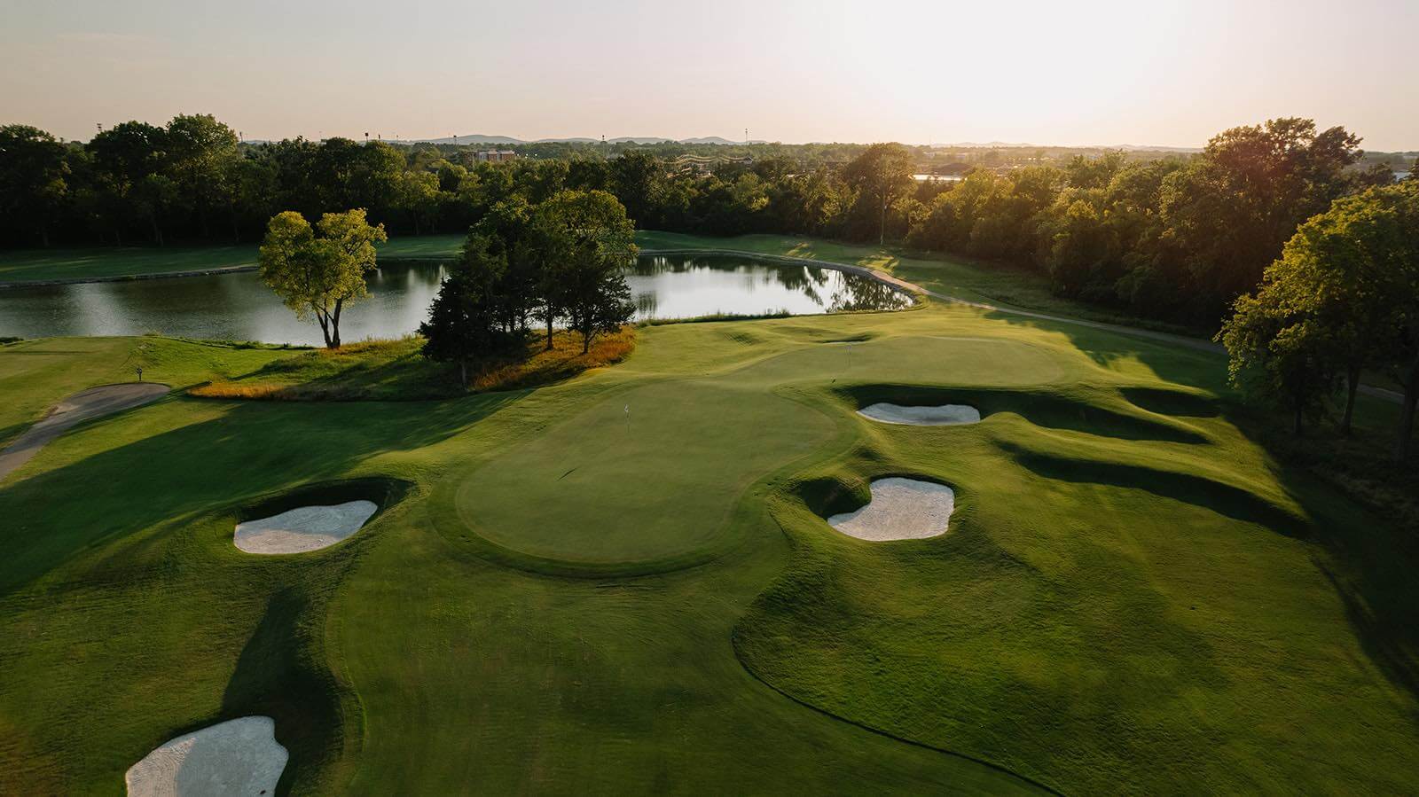 Aerial view of a Nashville golf course with sand bunkers, green fairways, a central water hazard, and surrounding trees at sunset.