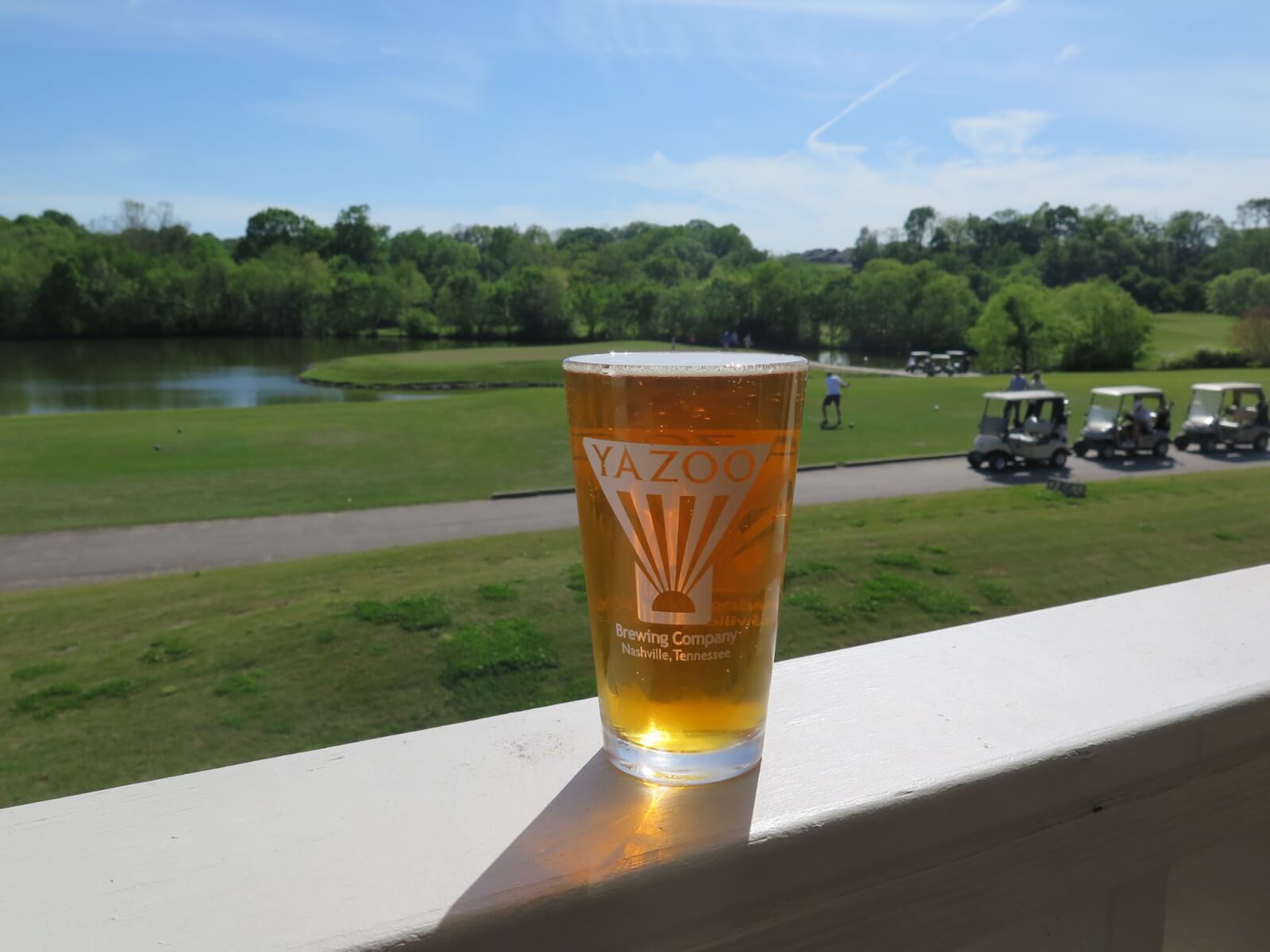 A pint glass of Yazoo beer sits on a white railing overlooking one of the scenic Nashville golf courses, with golf carts, a pond, and trees in the background.
