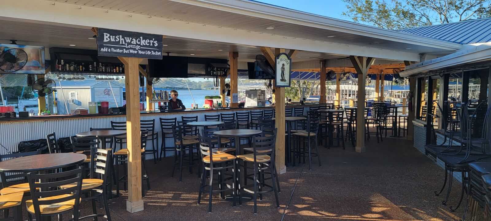 Your Guide to Old Hickory Lake - 6 Outdoor bar and dining area with high tables and chairs, a bartender behind the counter, and a scenic view of Old Hickory Lake in the background—perfect for enjoying lake activities from the Summer Guide.