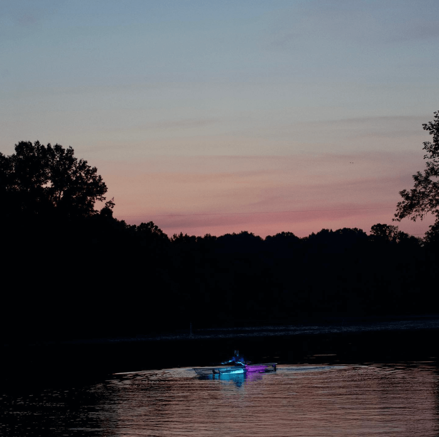 Your Guide to Old Hickory Lake - 2 A person in a kayak with blue and purple lights paddles on Old Hickory Lake at dusk, trees silhouetted against a pink and purple sky—an unforgettable scene for any Summer Guide.
