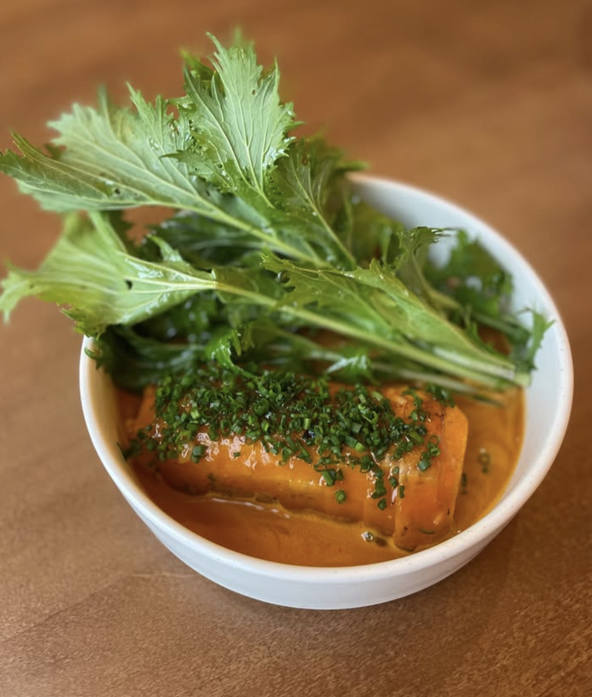 A bowl from Upscale Vegan Nashville featuring a piece of plant-based fish in orange sauce, topped with chopped chives and garnished with leafy greens.