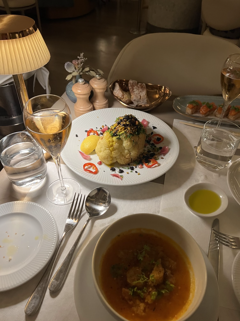 A restaurant table set with a bowl of soup, a roasted cauliflower dish, bread, wine glasses, a lamp, and various condiments showcases the charm of upscale plant-based meals found at top vegan restaurants in Nashville.