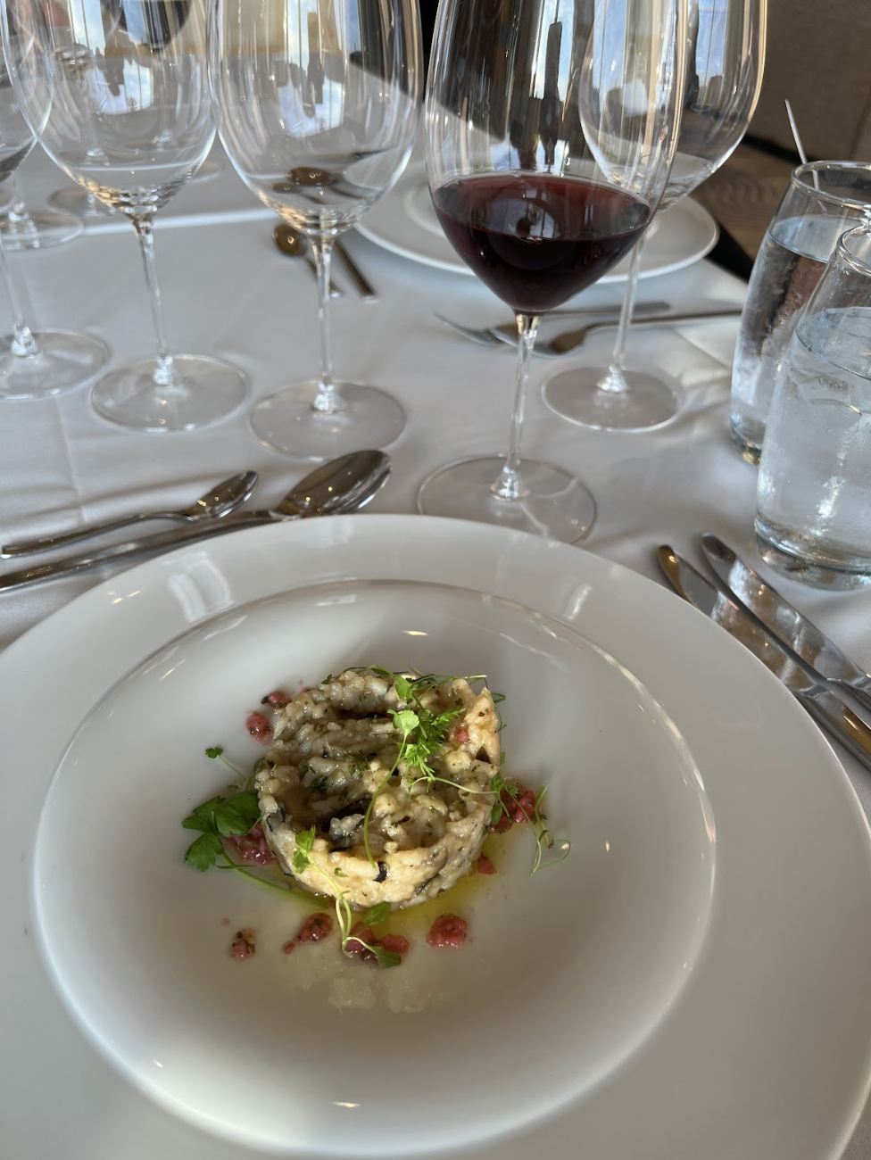 A plated gourmet vegan appetizer garnished with herbs is set on a white tablecloth next to a glass of red wine, water, and multiple empty wine glasses.
