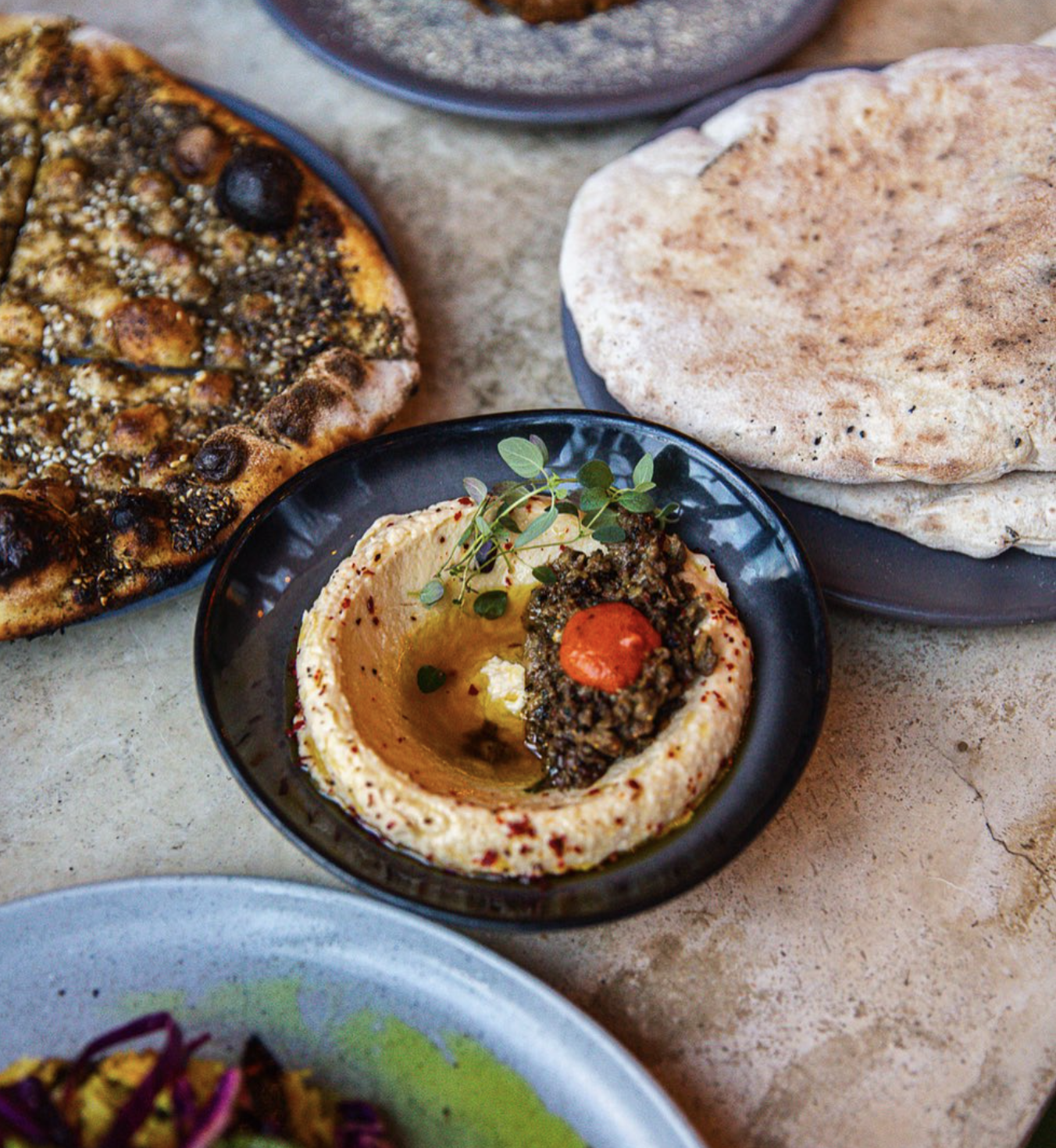 An upscale bowl of hummus topped with herbs, olive oil, and a red garnish sits next to flatbreads and za'atar bread.