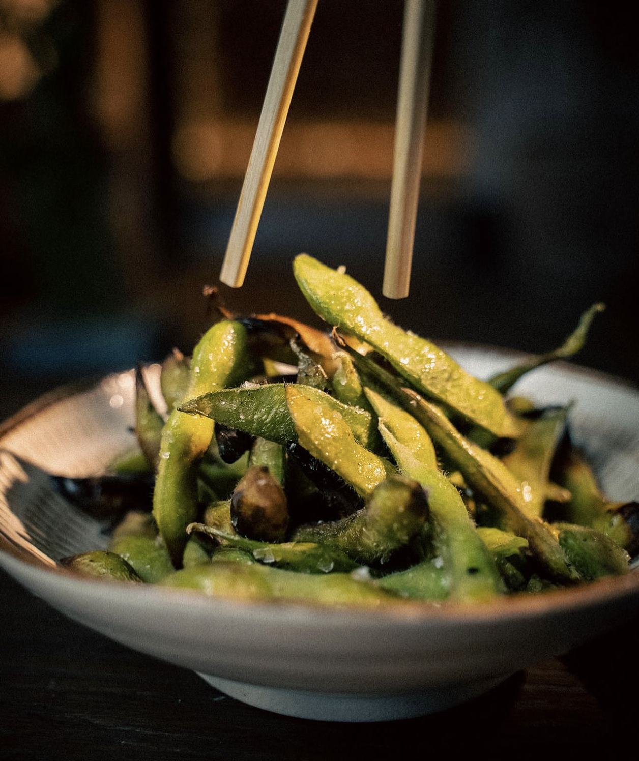 A bowl of grilled edamame pods, with a pair of chopsticks picking up one pod—an inviting bite from one of Nashville’s favorite vegan restaurants.