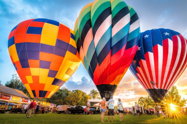 Three large, colorful hot air balloons are tethered on a grassy field at sunset during the Natchez Balloon Festival, with people standing nearby and tents in the background.