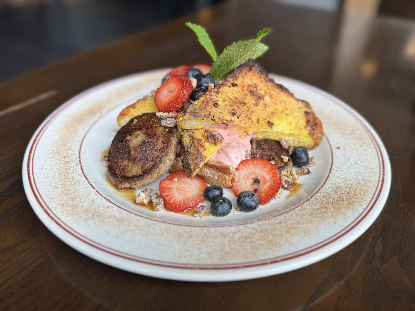 A plate of French toast topped with strawberries, blueberries, pecans, whipped pink cream, and a mint leaf, served with sausage on a white plate—discover this delicious Nashville Steals breakfast special in August 2025.