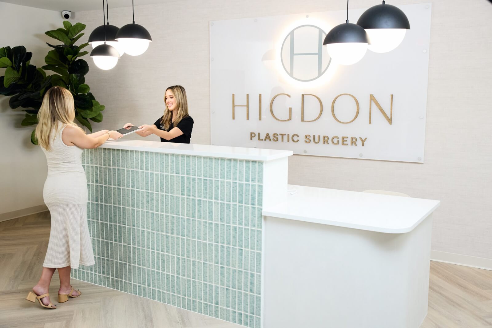 A woman stands at the reception desk of Higdon Plastic Surgery, handing paperwork to a receptionist under modern pendant lights—taking advantage of exclusive Nashville Deals available through August 2025.