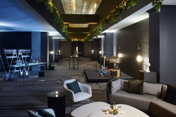 A modern lounge with sofas, armchairs, cocktail tables, a small stage with musical instruments, and overhead greenery decorations. Dim ambient lighting creates an intimate atmosphere—perfect for enjoying Nashville Deals in August 2025.