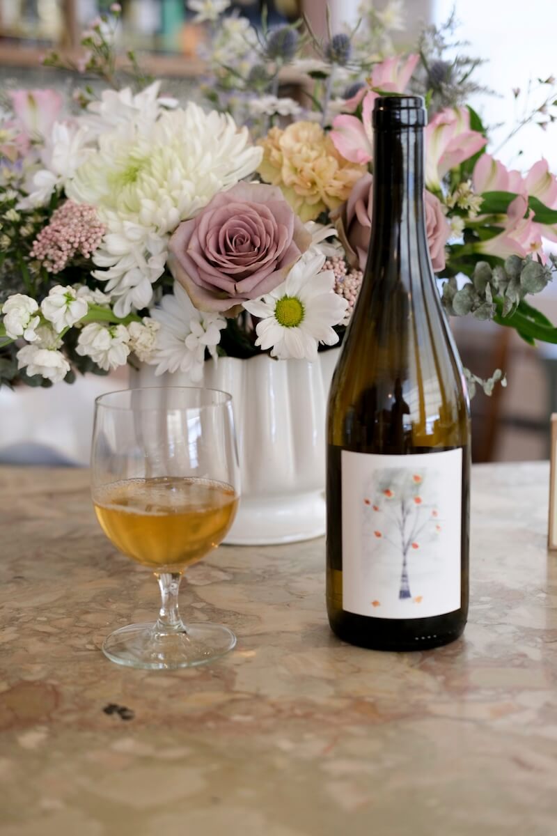 A glass of white wine and a wine bottle with an illustrated label sit on a marble surface next to a vase of assorted flowers—perfect summer wines to sip and savor on warm days.