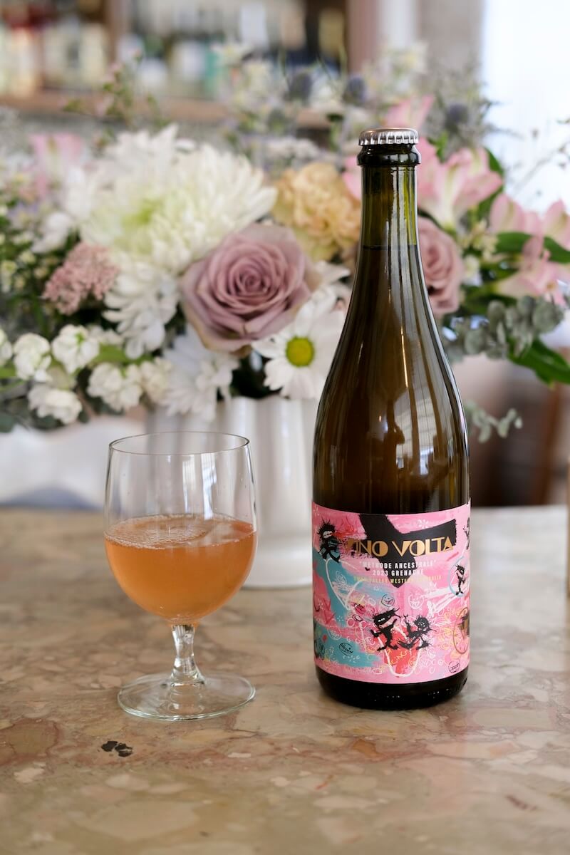 A glass of rosé-colored sparkling wine next to a bottle labeled "Indi Volta," set on a marble table with a bouquet of flowers in the background—perfect among refreshing wines to sip on warm summer days.