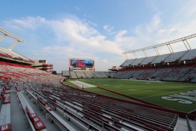 Wide view of an empty South Carolina football stadium with rows of red and white seats, a large scoreboard, and a green field under a partly cloudy sky.