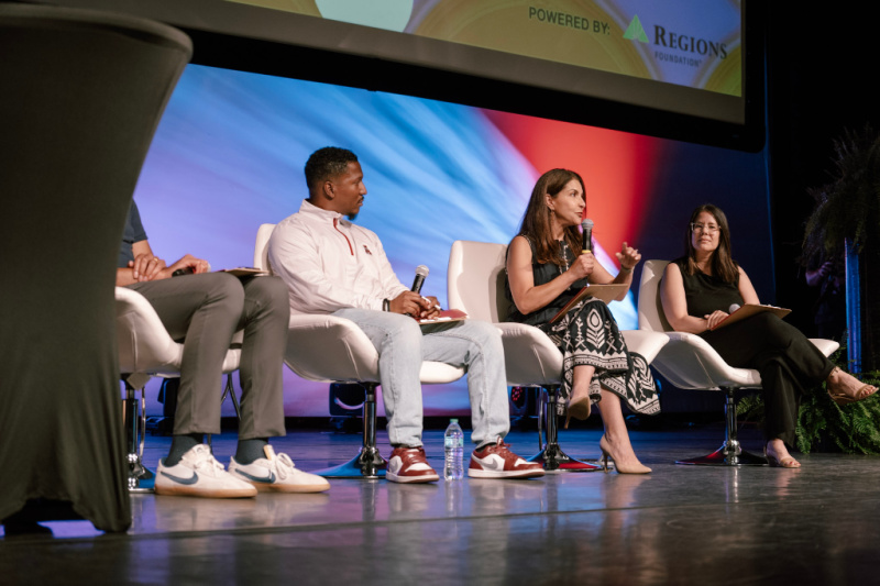 Four panelists sit on stage in white chairs, holding microphones and engaged in discussion; a screen and signage are visible in the background.