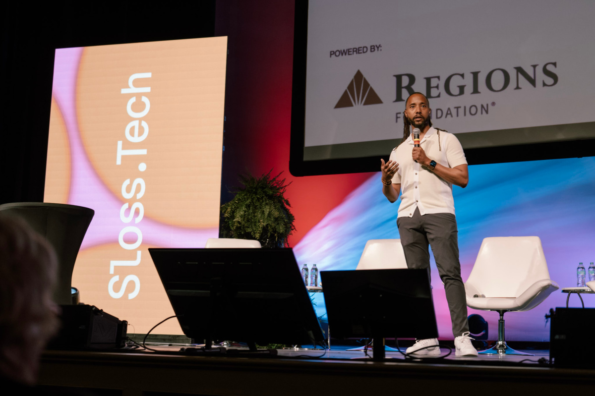A man stands on stage speaking at the Sloss Tech conference, with a large screen and event sponsor logos.