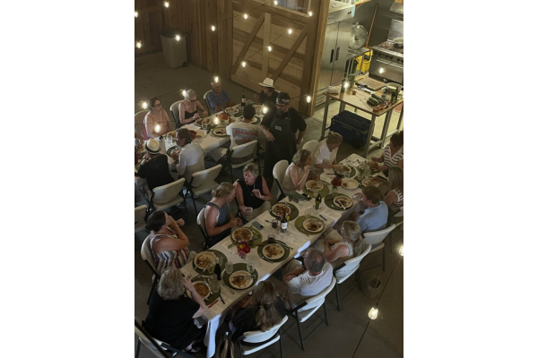 A group of people sit at two long tables enjoying a Sunday Supper together in a rustic, warmly lit indoor setting.