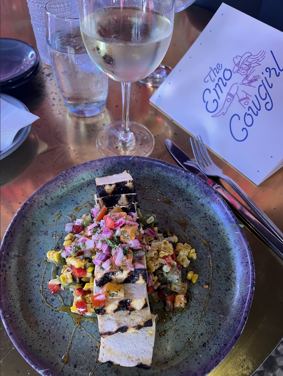 A plated dish of grilled halloumi with corn salsa sits on a green ceramic plate beside a glass of white wine and a menu reading "The Emo Cowgirl," highlighting the upscale vegan Nashville dining scene.