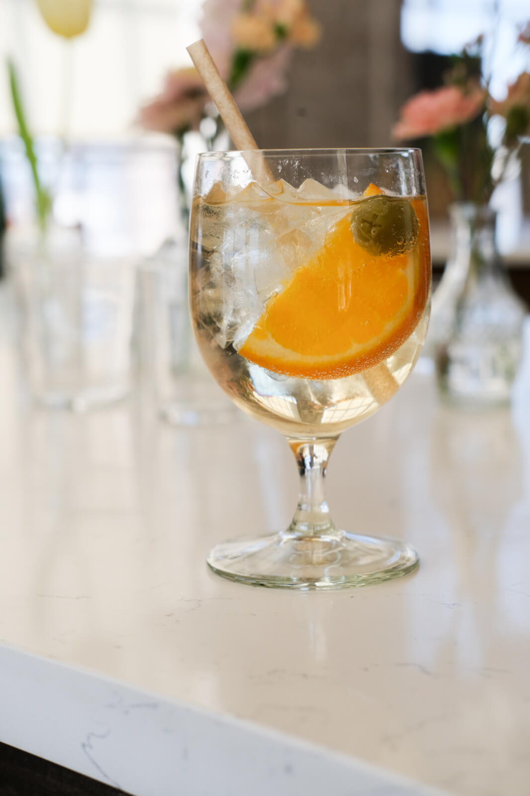 A stemmed glass with ice, an orange slice, and an olive sits on a white countertop, evoking the refreshing allure of a Paros Cocktail, with blurred flowers in the background.