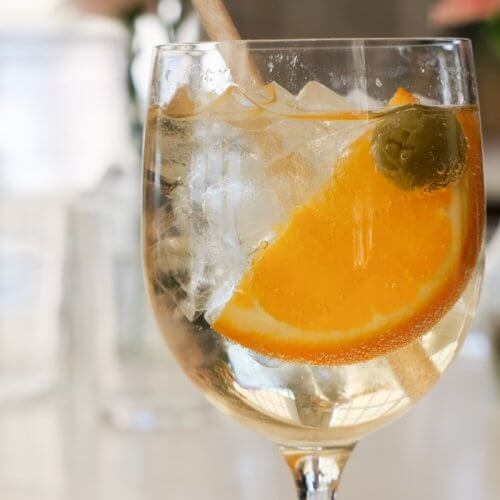 A glass of Paros Cocktail garnished with an orange slice and a green olive, with a straw, sits on a table; blurred glassware in the background hints at an unforgettable Night in Paros.