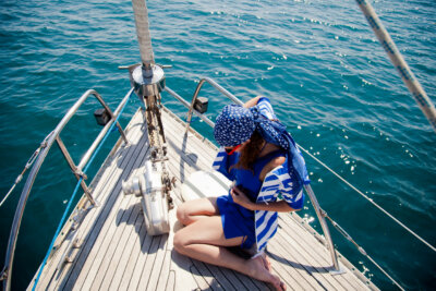 A person in a blue and white outfit demonstrates scarf tying with their headscarf as they sit on the deck of a sailboat, looking down, calm blue water in the background.