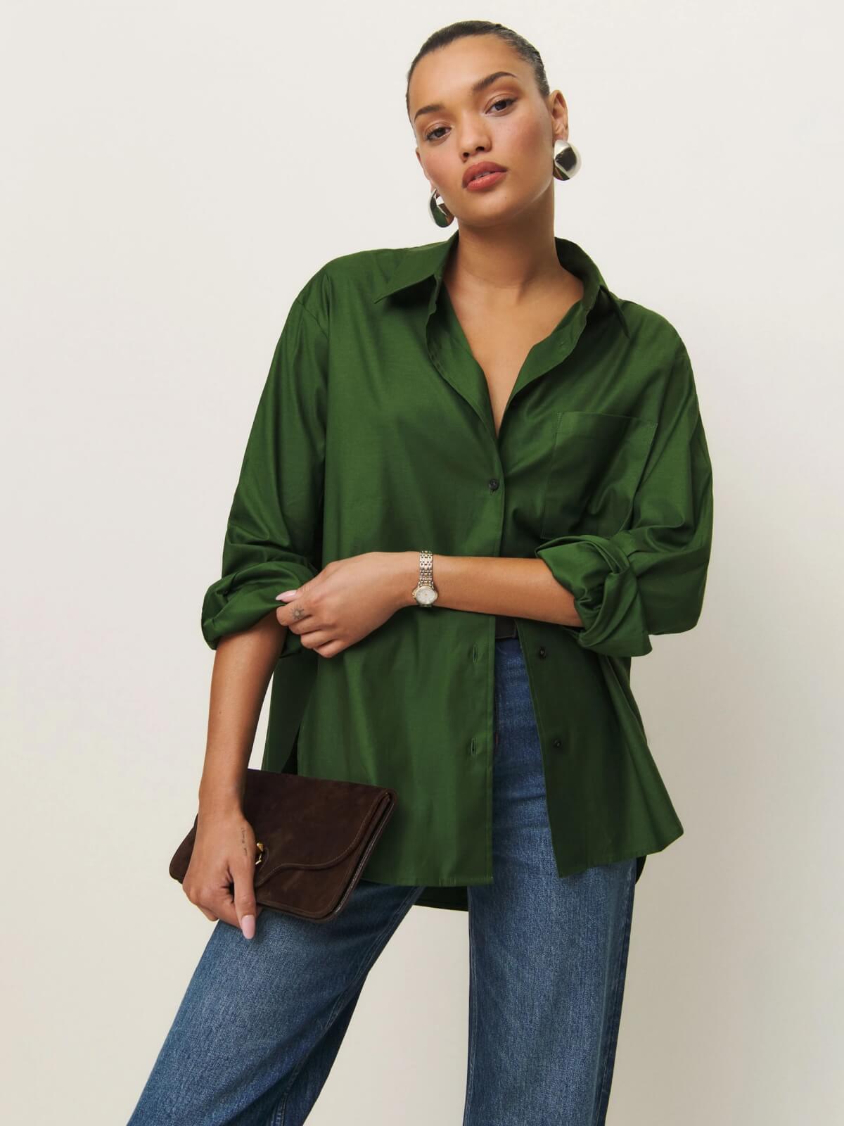 A woman wearing a loose green button-up shirt and blue jeans holds a brown clutch bag. She finishes the look with hoop earrings, a wristwatch, and her hair pulled back.