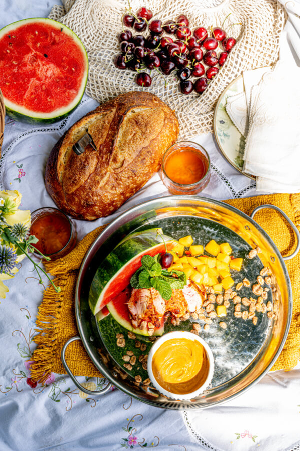 A Dame Fine Picnic spread with bread, watermelon slice, cherries, mango cubes, granola, a small dip, and two glasses of juice on a table with a yellow cloth and floral tablecloth.