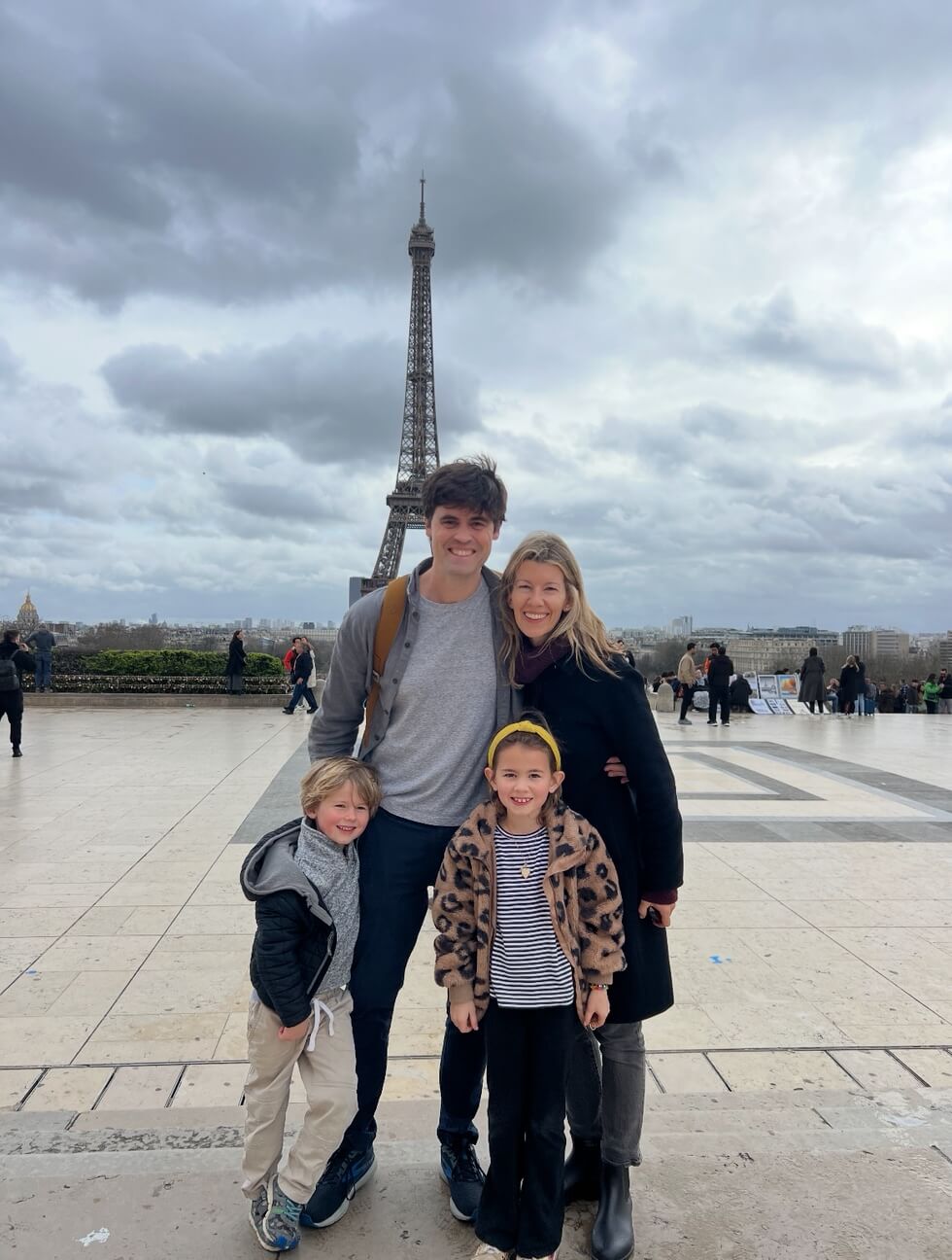 A family of four stands smiling in front of the Eiffel Tower in Paris on a cloudy day.