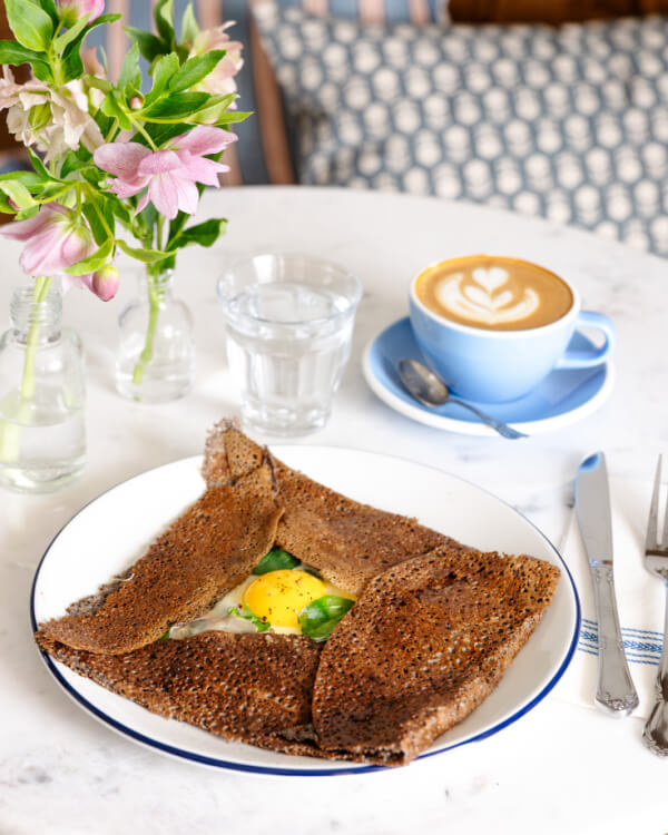 A buckwheat galette with egg and greens sits on a plate at The Yellow Table, accompanied by a latte, a glass of water, cutlery, and a vase with flowers atop the marble surface.