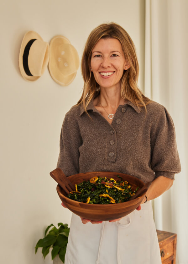 A woman holds a large wooden bowl of salad and smiles at the camera. Two hats hang on the wall in the background.
