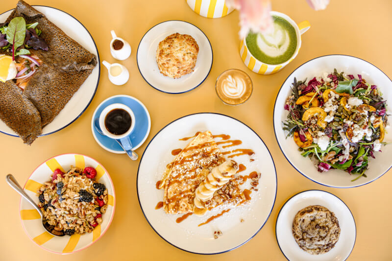 A table set with various breakfast dishes from The Yellow Table, including crepes, a salad, baked goods, parfait, a savory galette, and cups of coffee and matcha latte.