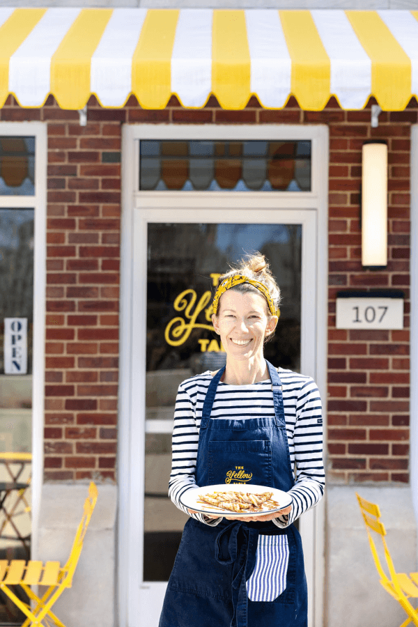 A woman in a striped shirt and apron stands outside The Yellow Table café, holding a plate of food, with yellow chairs and a striped awning in the background.