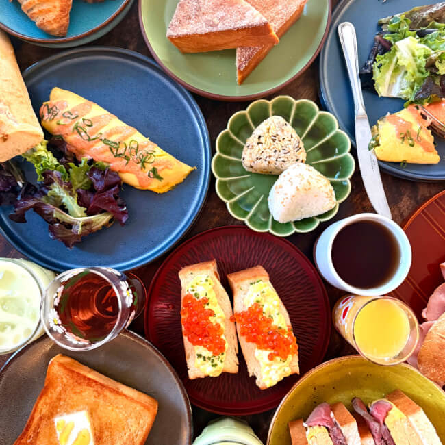A table set with various breakfast foods—from omelets to rice balls and toast. Beverages and assorted side dishes complete this tempting morning spread.