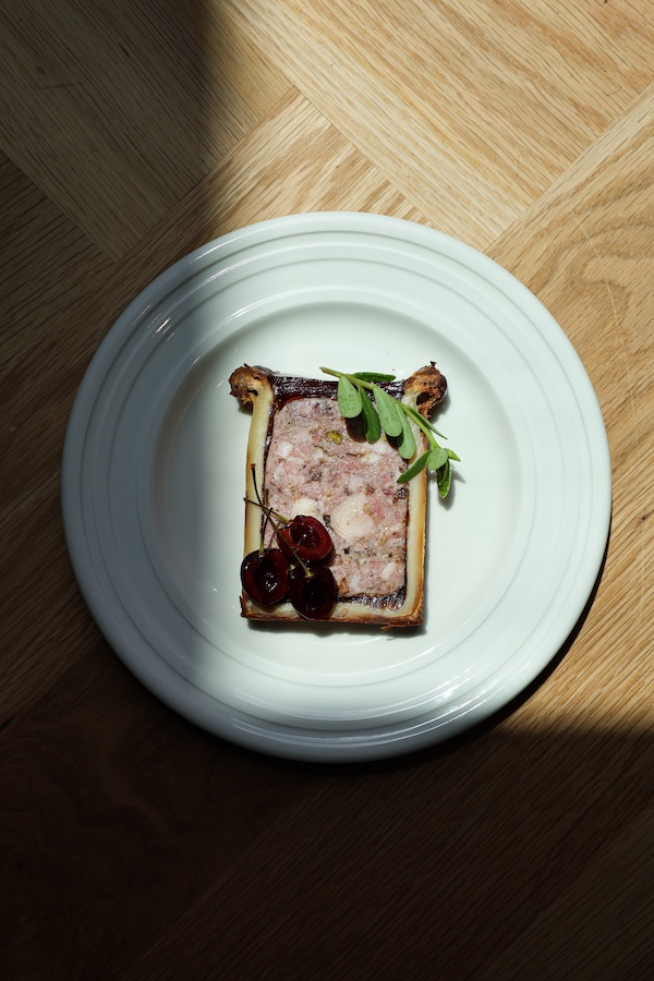 A slice of pâté en croûte is served on a white plate, garnished with herbs and cherries, on a wooden surface with a shadow cast across part of the scene—a tempting dish found at new Nashville restaurants.