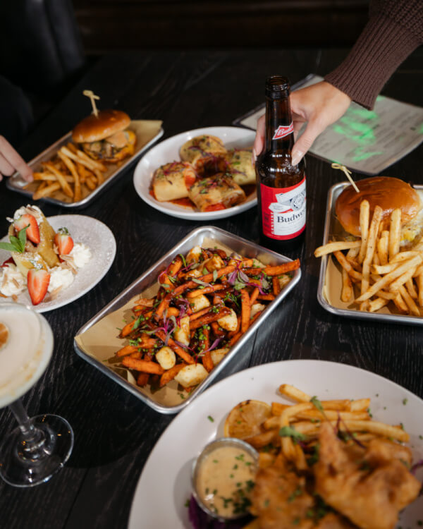A table with plates of burgers, fries, sweet potato fries, bruschetta, a salad, and a hand reaching for a bottle of Budweiser beer captures the lively spirit of Nashville restaurants this July 2025.