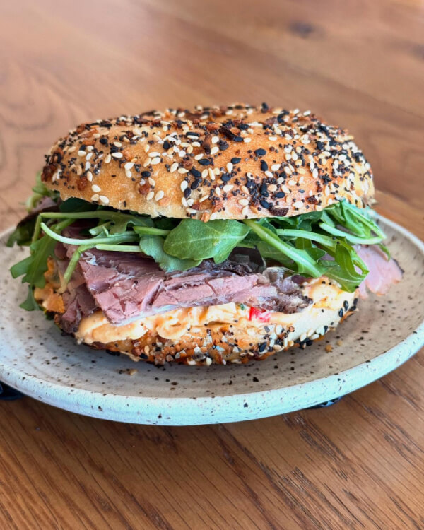 A sesame seed bagel sandwich with roast beef, arugula, and a creamy spread, served on a ceramic plate.