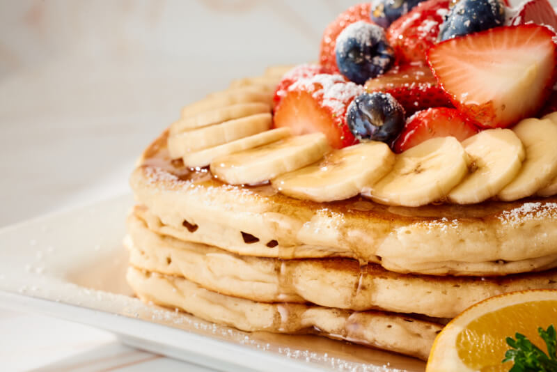 Stack of pancakes topped with banana slices, strawberries, blueberries, powdered sugar, and syrup, with a lemon wedge on the side.