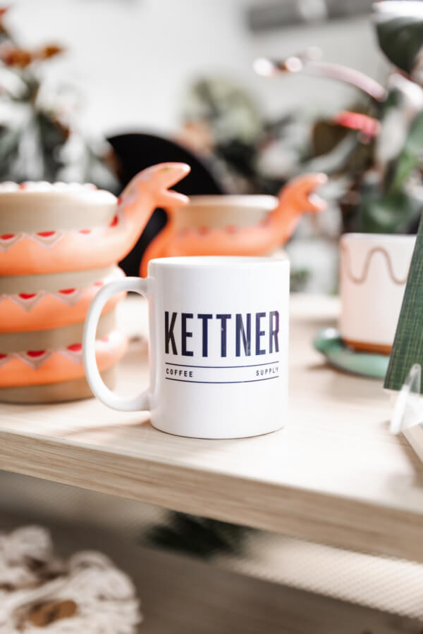 A white mug with the text "KETTNER COFFEE SUPPLY" sits on a wooden surface, with decorative teapots and plants in the background.
