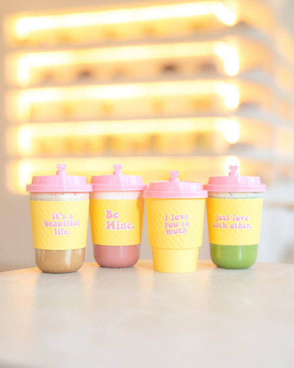 Four colorful drinks in cups with pink lids and yellow sleeves, each featuring a positive phrase, on a light table with a blurred background.