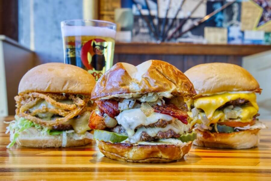 Three cheeseburgers with various toppings are arranged side by side on a wooden table; a glass of beer is in the background at one of the hottest new restaurants in Nashville, opening July 2025.