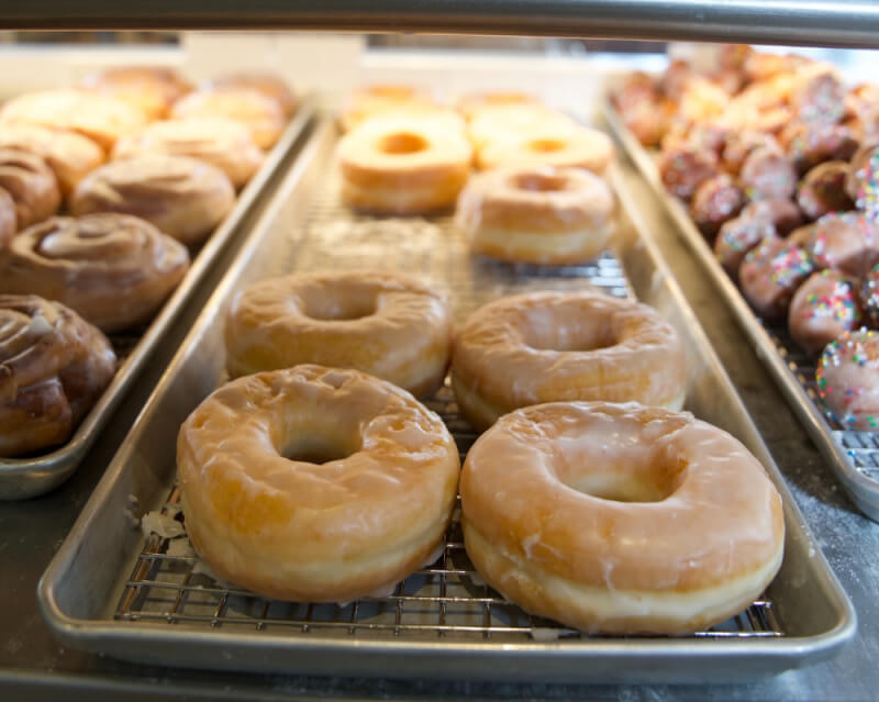 Trays of glazed donuts, cinnamon rolls, and sprinkled donuts are displayed on metal racks in a bakery case.