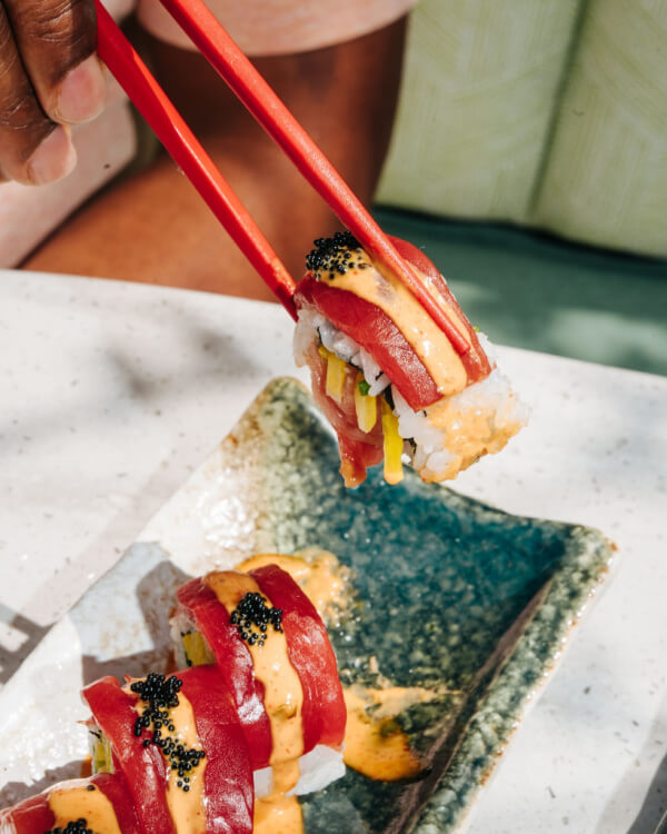 A hand uses red chopsticks to pick up a sushi roll topped with tuna, orange sauce, and black roe from a rectangular ceramic plate at one of the exciting new Nashville restaurants opening July 2025.