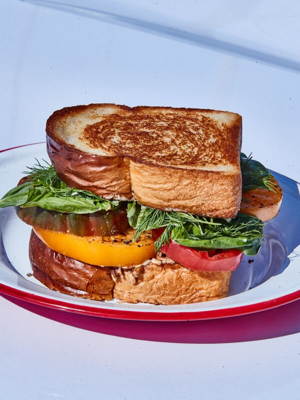 A delicious tomato dish, this grilled sandwich features thick slices of bread, yellow and red tomatoes, leafy greens, and herbs served on a white plate with a red rim.