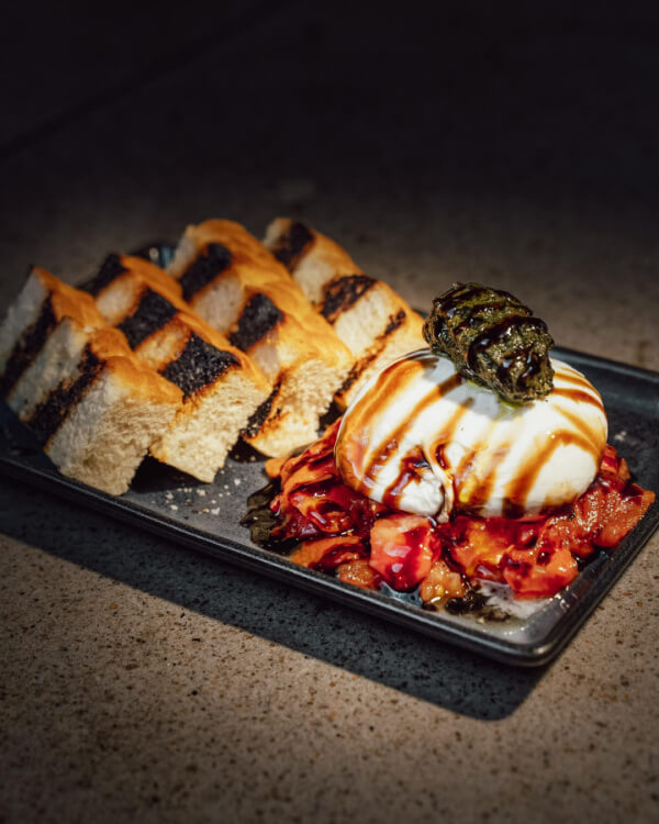 A rectangular plate with grilled bread slices, burrata cheese drizzled with balsamic glaze, roasted vegetables, and pesto on top—perfect for fans of flavorful tomato dishes.