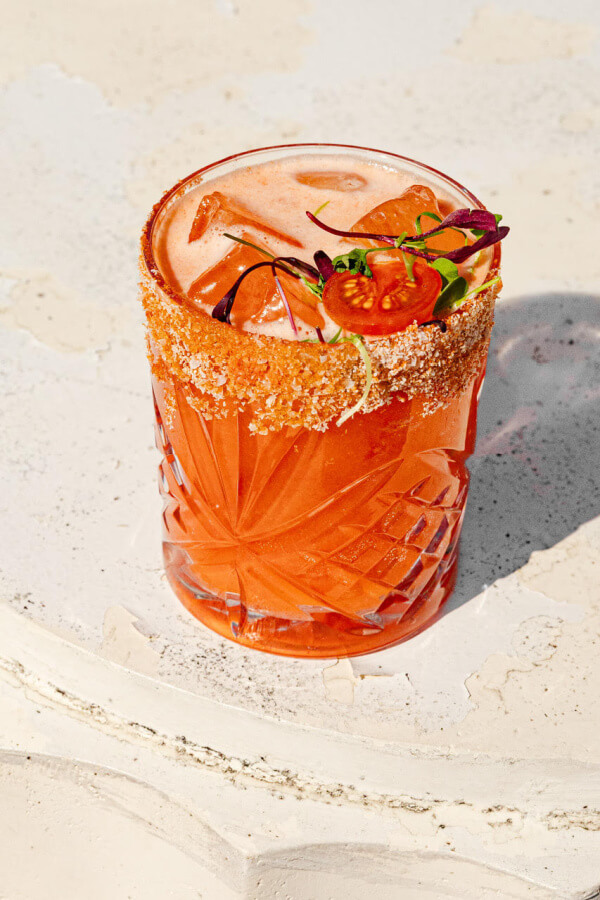 A glass of bright orange cocktail with a chili-salt rim, ice, and garnished with tomato and microgreens—perfect for fans of creative tomato dishes—set on a textured white surface.