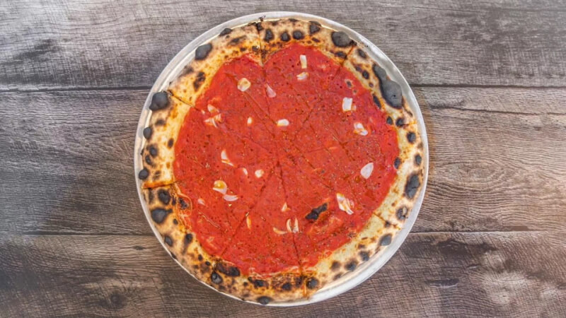 A whole pizza with a thin, charred crust, tomato sauce, and garlic slices—a classic among tomato dishes—served on a metal tray atop a wooden table.