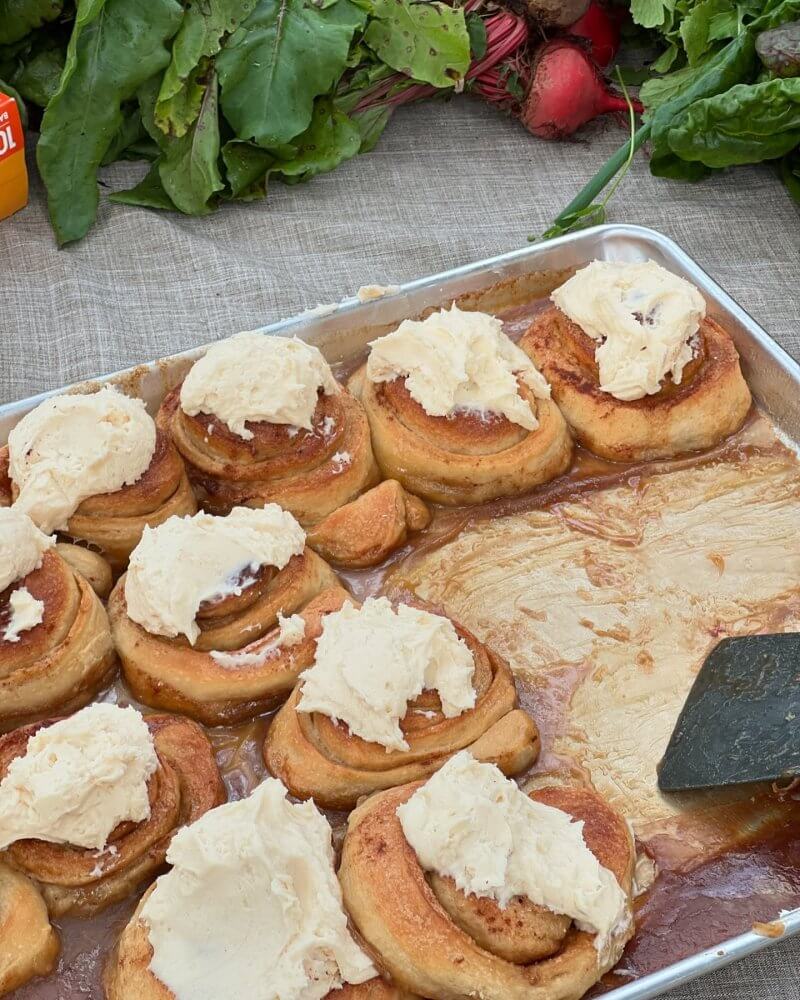 A tray of cinnamon rolls topped with frosting sits on a table near leafy greens, capturing the local flavor and coastal charm often found at 30A farmers markets, with a spatula resting on the tray.