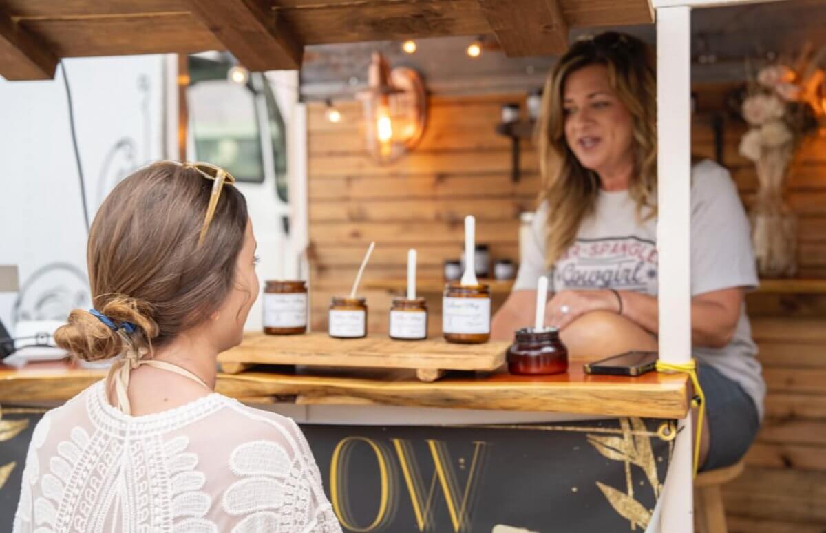A woman behind a wooden counter displays jars of honey to another woman at an outdoor market stall, capturing the coastal charm and local flavor of 30A farmers markets.
