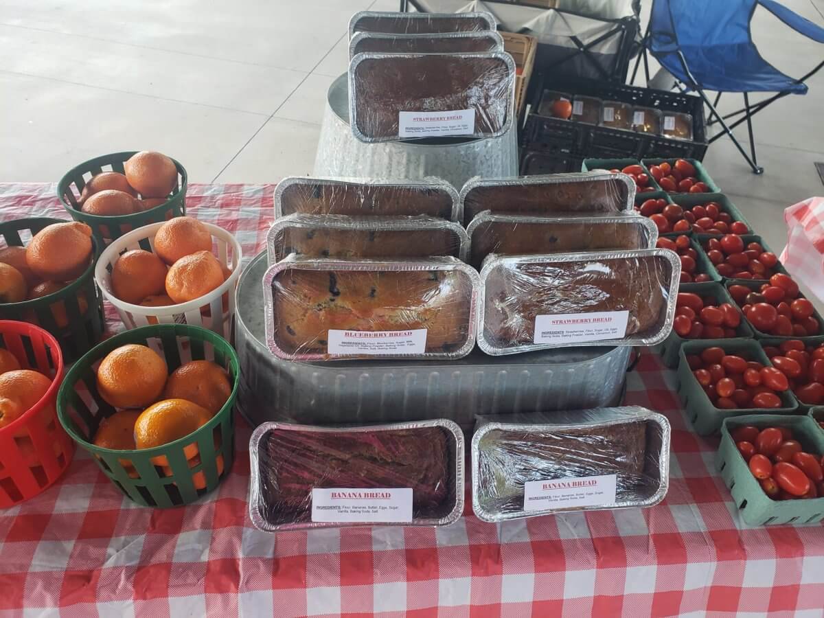 Loaves of homemade bread—zucchini, strawberry, and banana—are displayed on a table with baskets of oranges and cherry tomatoes, capturing the fresh finds and local flavor of 30A farmers markets.
