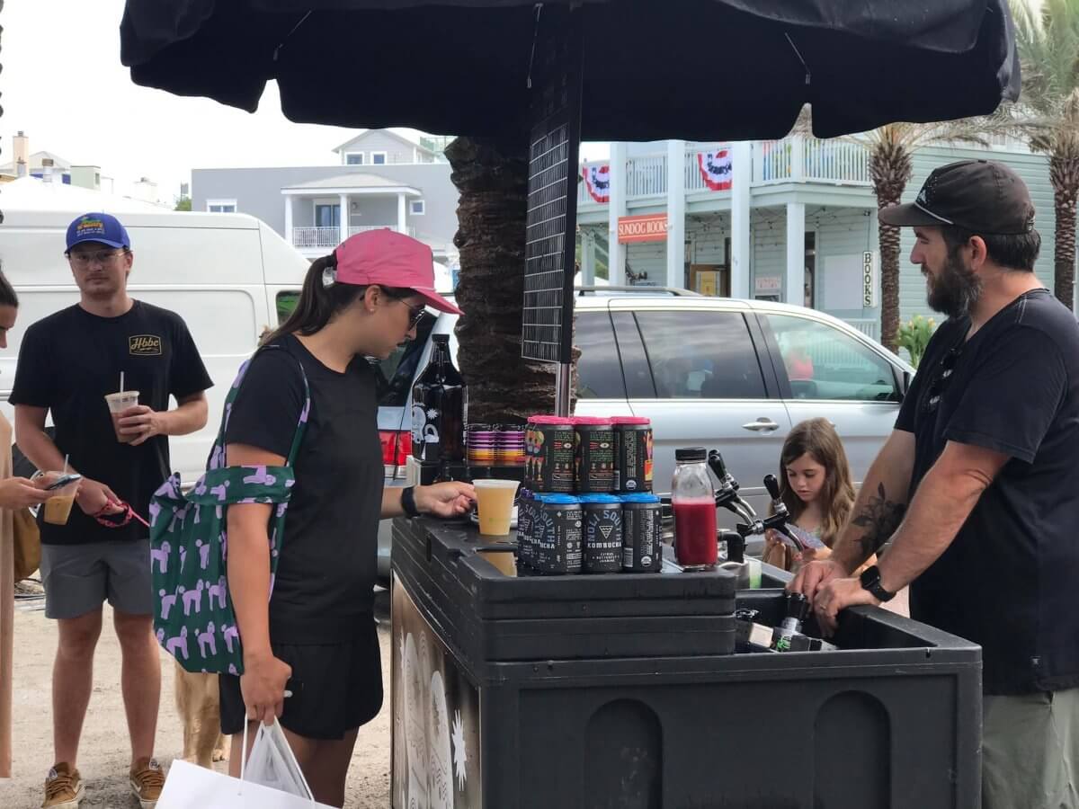 A woman orders a drink from a vendor at an outdoor stand, capturing the local flavor of 30A farmers markets; others wait nearby while houses and a parked van appear in the background.